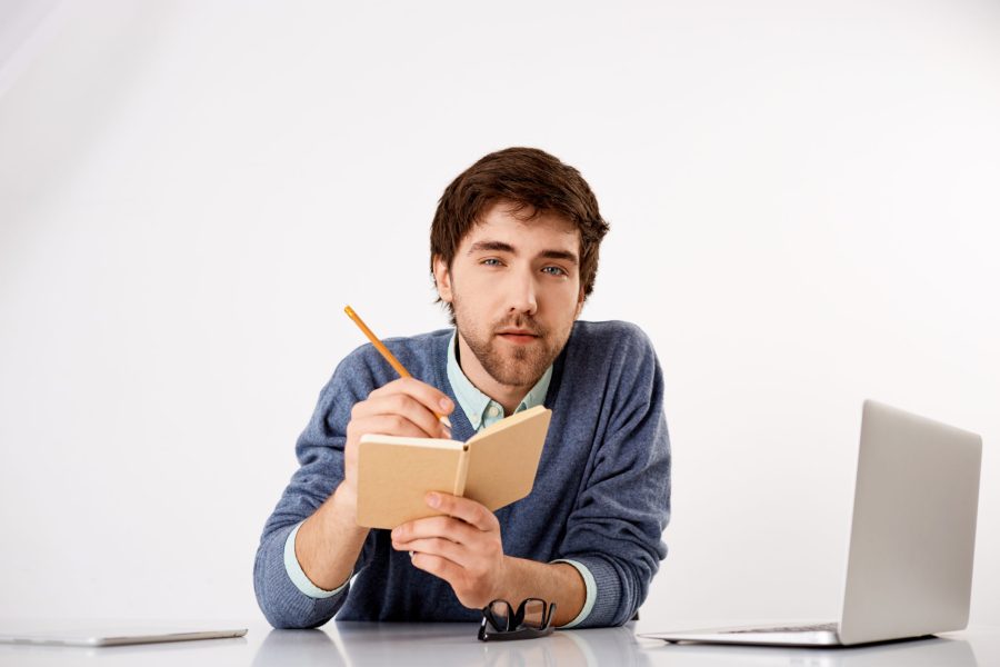 Handsome confident concentrated young businessman sitting at the office desk writing down something to notebook with a pencil. Looking at camera. Isolated on white background.